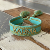 Close-up of adjustable turquoise woven bracelet with gold embroidered “KARMA” lettering and sliding knot tassel cords.