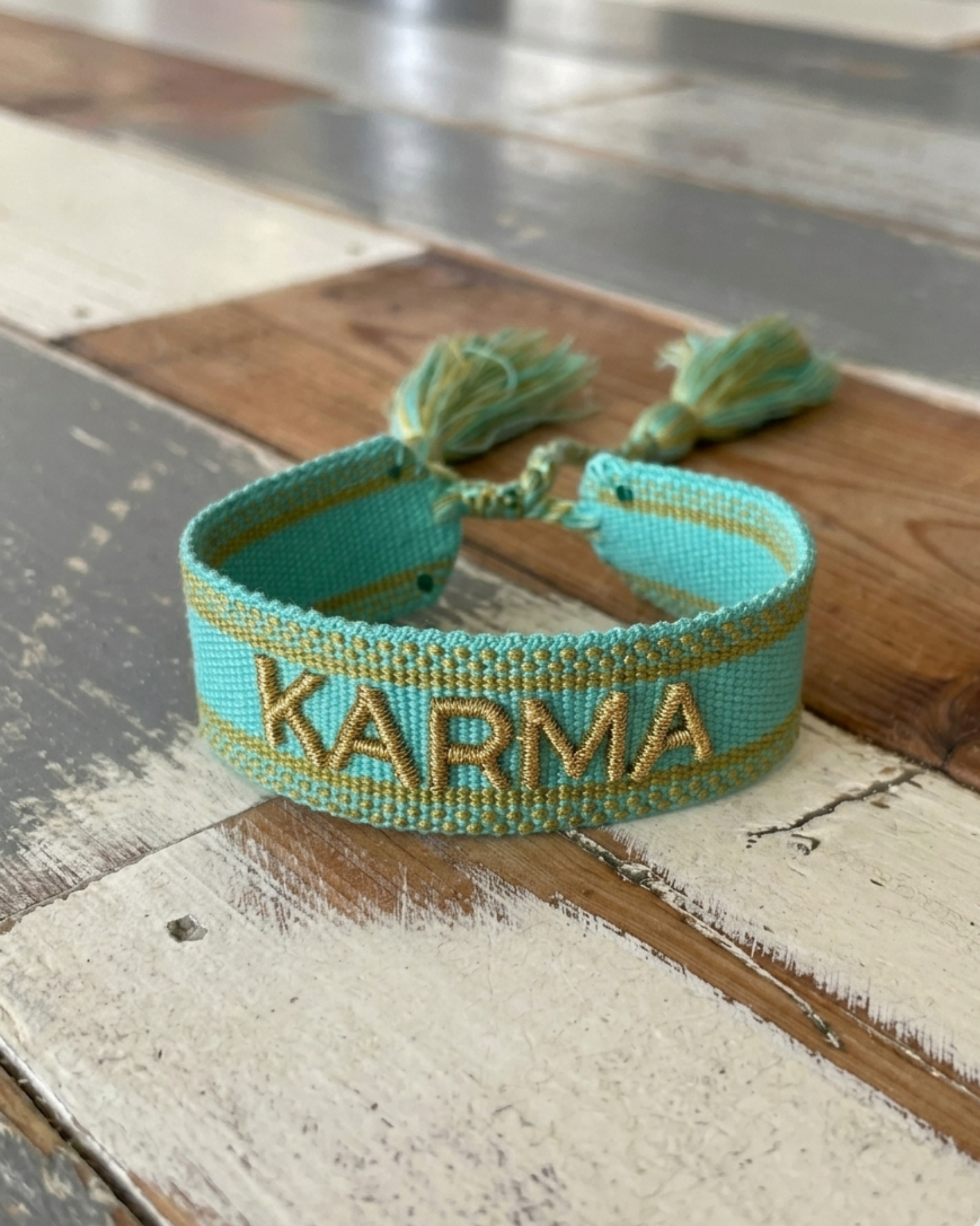 Close-up of adjustable turquoise woven bracelet with gold embroidered “KARMA” lettering and sliding knot tassel cords.
