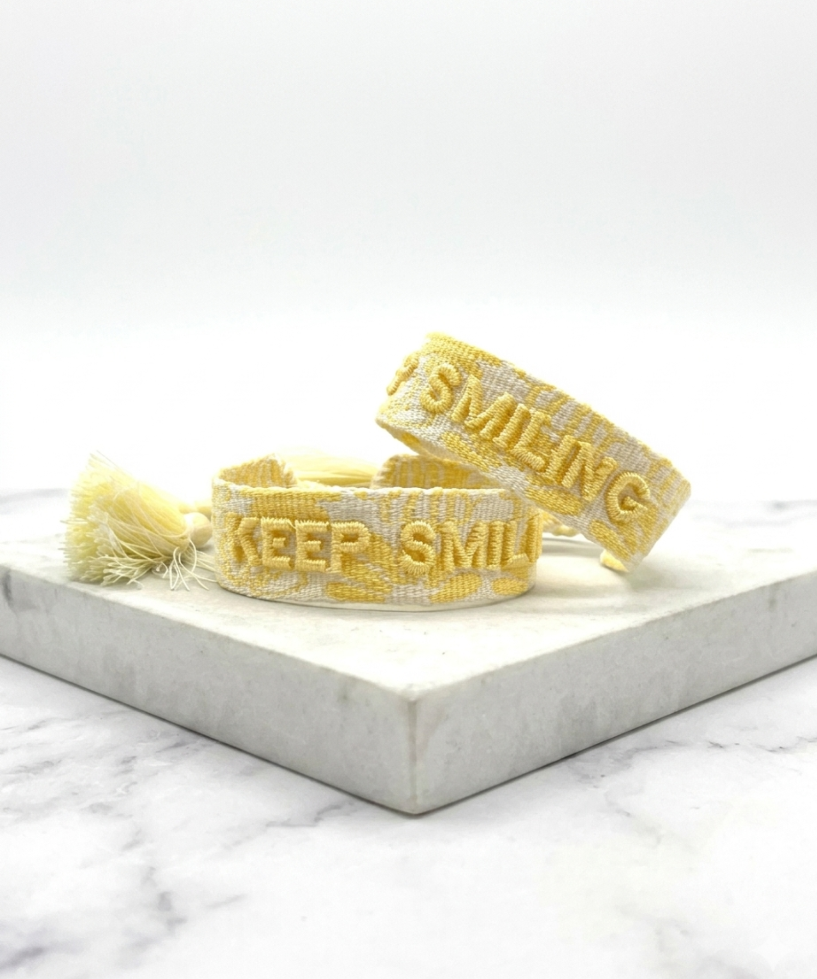 Close-up of adjustable yellow sorbet woven bracelet with embroidered “KEEP SMILING” lettering and sliding knot tassel cords.
