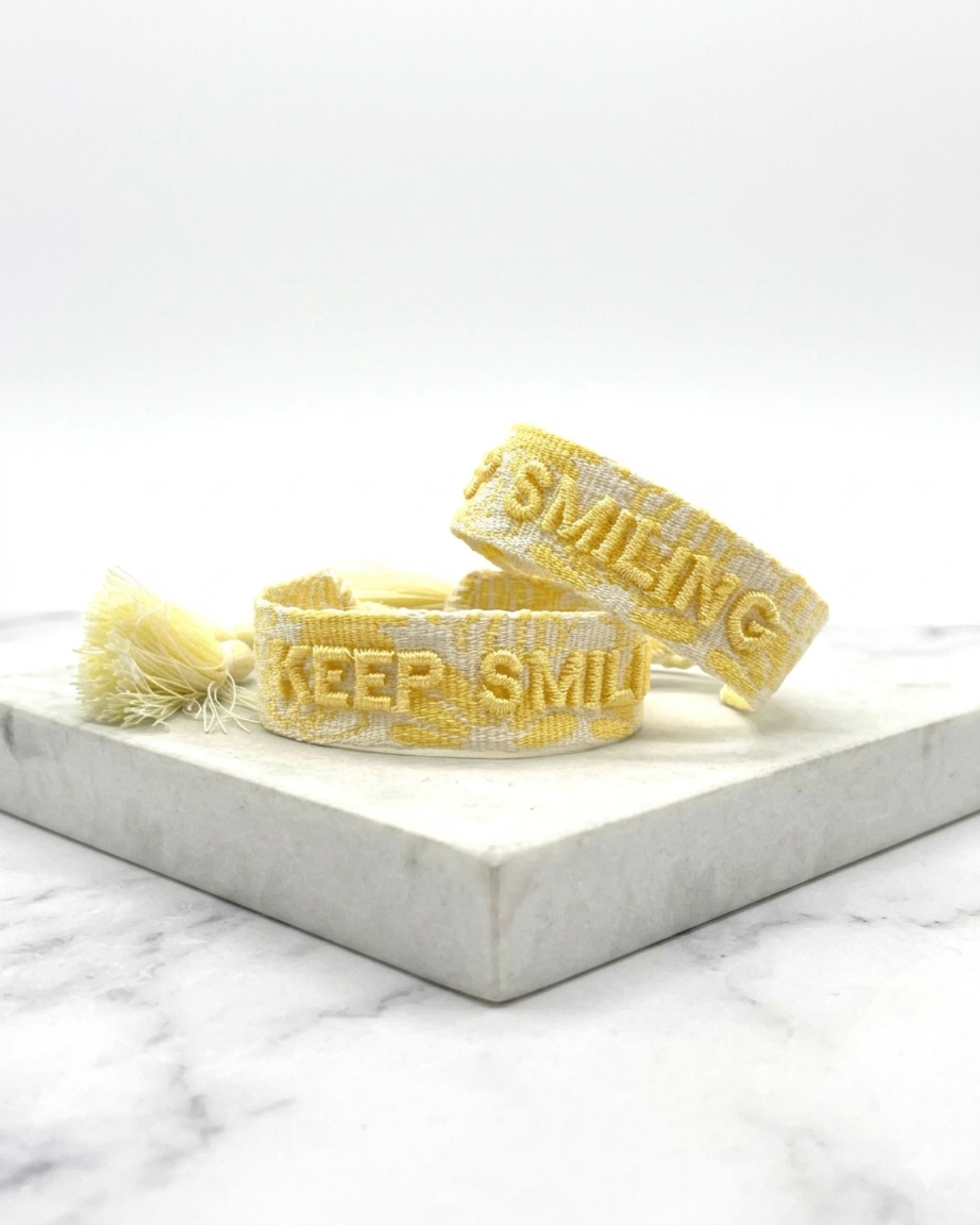 Close-up of adjustable yellow sorbet woven bracelet with embroidered “KEEP SMILING” lettering and sliding knot tassel cords.