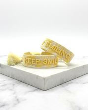 Close-up of adjustable yellow sorbet woven bracelet with embroidered “KEEP SMILING” lettering and sliding knot tassel cords.