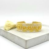Close-up of adjustable yellow sorbet woven bracelet with embroidered “KEEP SMILING” lettering and sliding knot tassel cords.