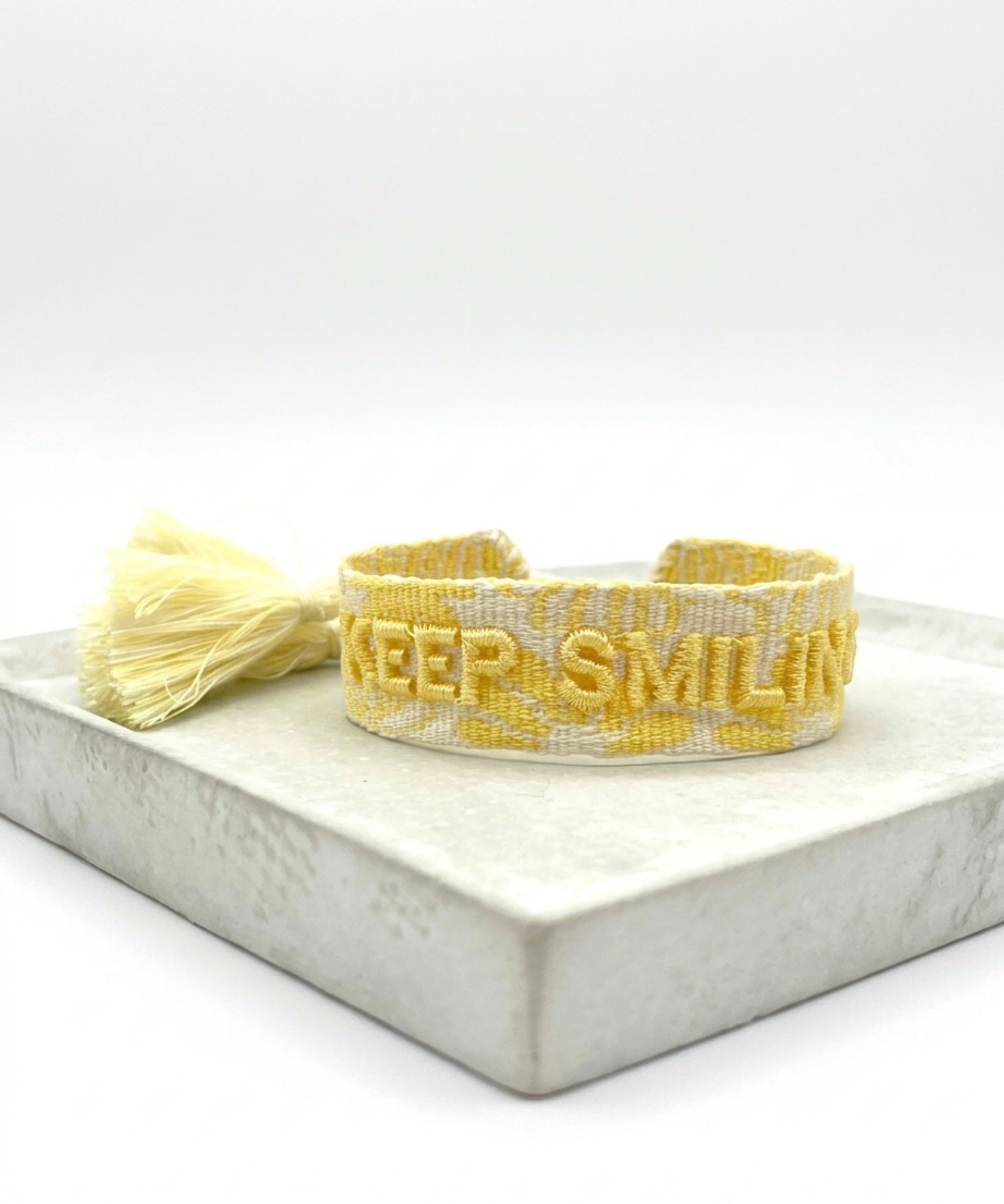 Close-up of adjustable yellow sorbet woven bracelet with embroidered “KEEP SMILING” lettering and sliding knot tassel cords.