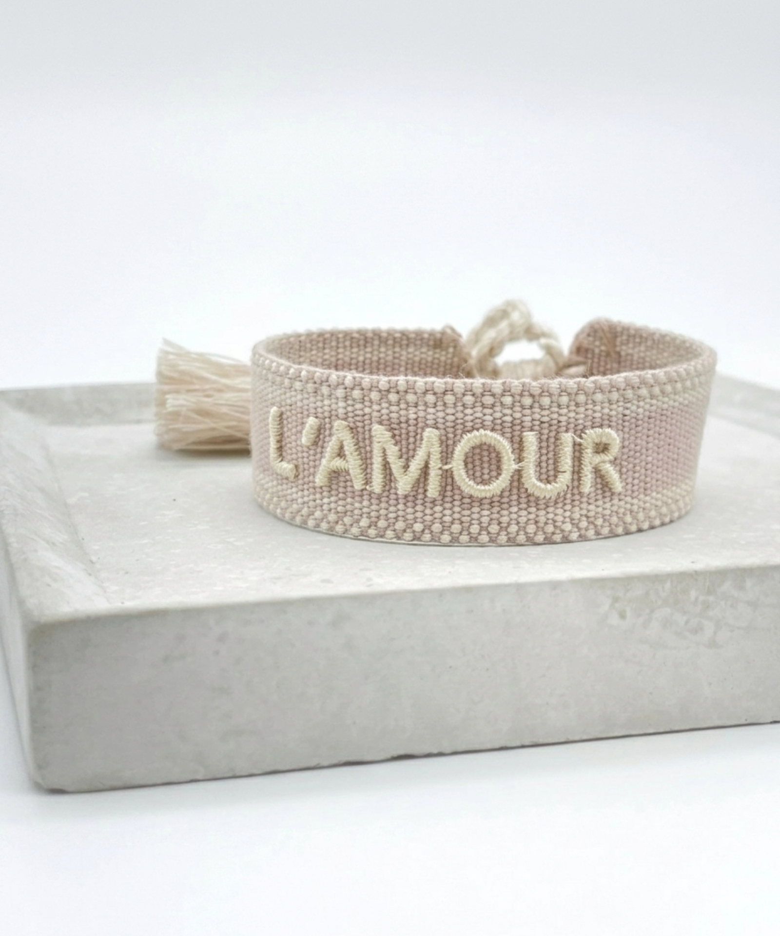 Close-up of adjustable light taupe woven bracelet with ecru embroidered “L'AMOUR” lettering, matching border, and sliding knot tassel cords.
