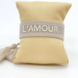 Close-up of adjustable light taupe woven bracelet with ecru embroidered “L'AMOUR” lettering, matching border, and sliding knot tassel cords.