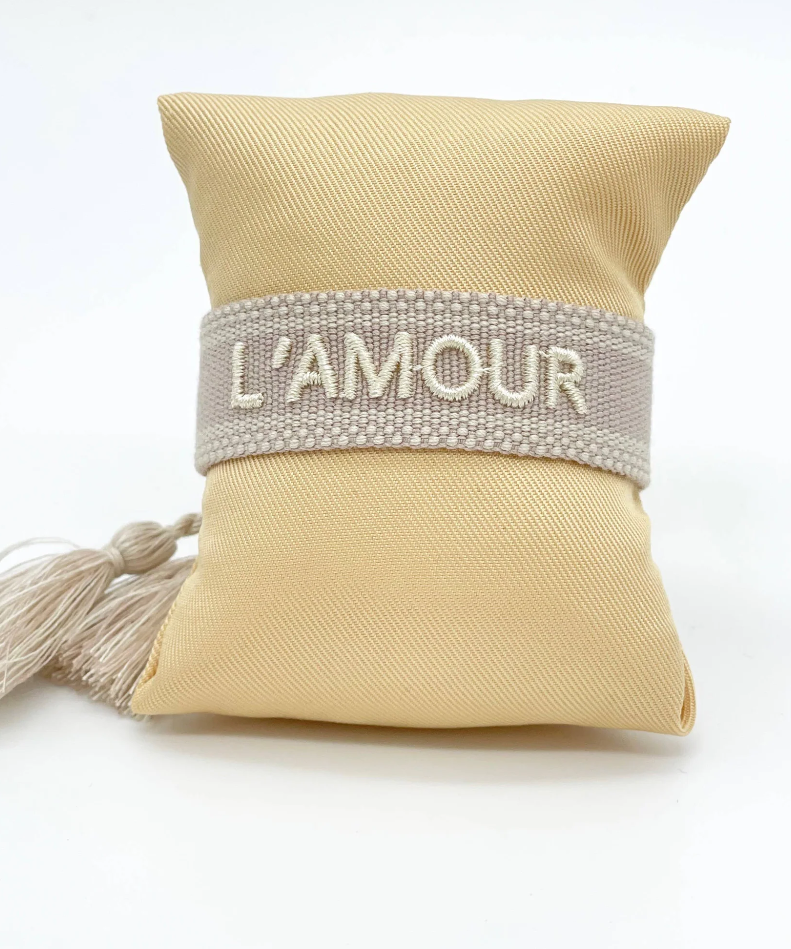 Close-up of adjustable light taupe woven bracelet with ecru embroidered “L'AMOUR” lettering, matching border, and sliding knot tassel cords.