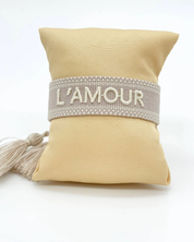 Close-up of adjustable light taupe woven bracelet with ecru embroidered “L'AMOUR” lettering, matching border, and sliding knot tassel cords.