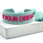 Close-up of adjustable sage green woven friendship bracelet with dark pink embroidered "LIVE YOUR DREAMS" lettering and sliding knot cords.