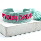 Close-up of adjustable sage green woven friendship bracelet with dark pink embroidered "LIVE YOUR DREAMS" lettering and sliding knot cords.