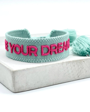 Close-up of adjustable sage green woven friendship bracelet with dark pink embroidered "LIVE YOUR DREAMS" lettering and sliding knot cords.