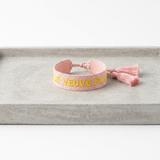 Close-up of adjustable salmon woven boho bracelet with offwhite border and orange embroidered “MORE VEUVE PLEASE” lettering, featuring sliding knot tassel cords.