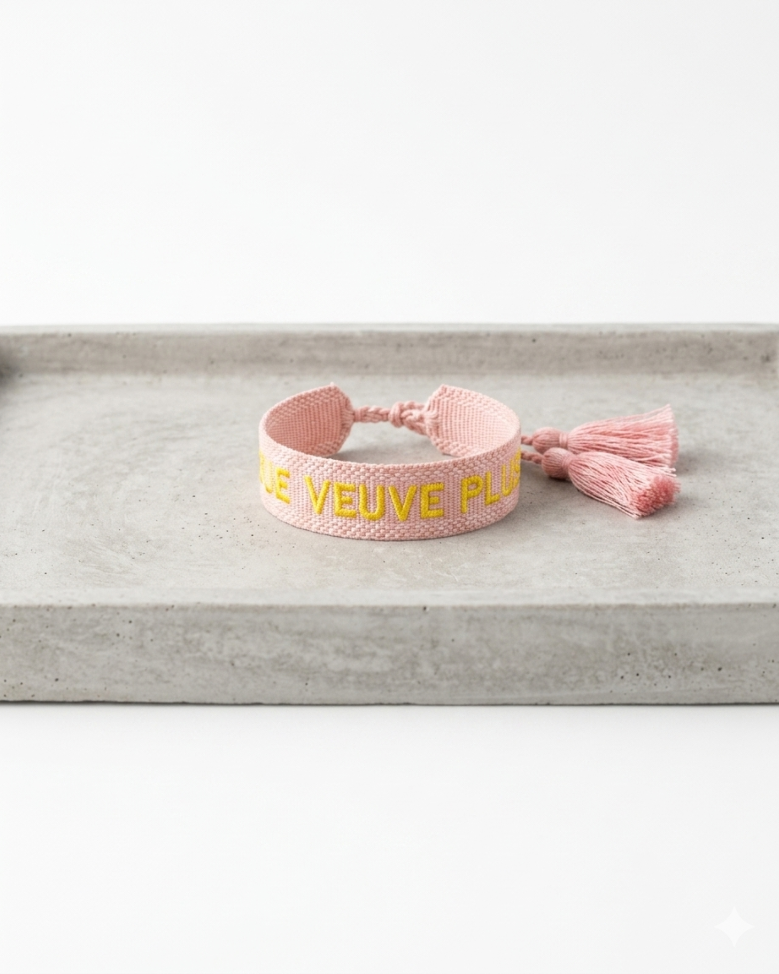 Close-up of adjustable salmon woven boho bracelet with offwhite border and orange embroidered “MORE VEUVE PLEASE” lettering, featuring sliding knot tassel cords.