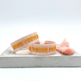Close-up of adjustable salmon woven boho bracelet with offwhite border and orange embroidered “MORE VEUVE PLEASE” lettering, featuring sliding knot tassel cords.