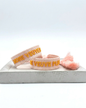 Close-up of adjustable salmon woven boho bracelet with offwhite border and orange embroidered “MORE VEUVE PLEASE” lettering, featuring sliding knot tassel cords.