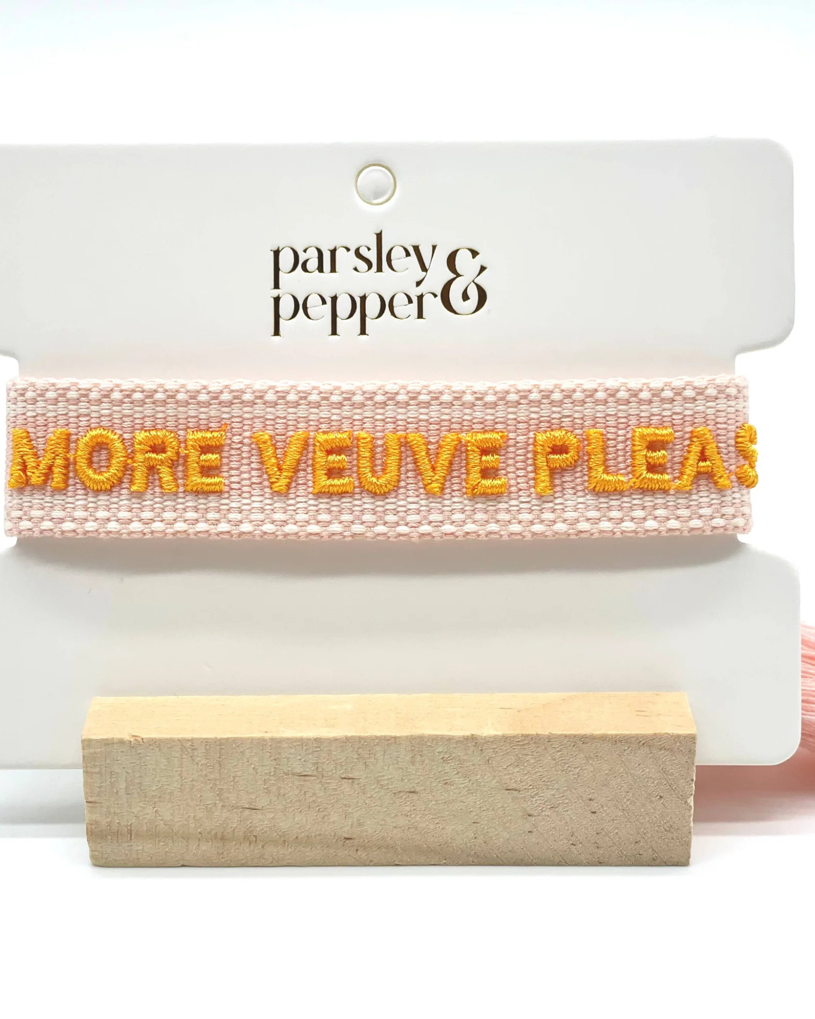 Close-up of adjustable salmon woven boho bracelet with offwhite border and orange embroidered “MORE VEUVE PLEASE” lettering, featuring sliding knot tassel cords.
