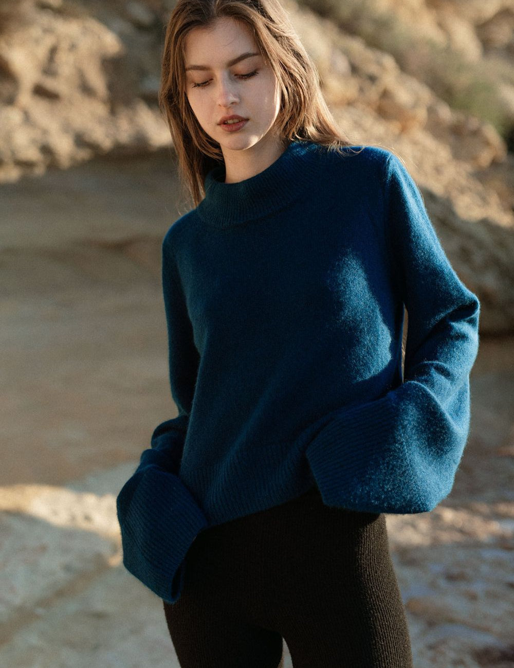 Woman wearing a petrol blue cashmere sweater by Nuage Cashmere on the beach, showing the front with wide sleeves and a high neck