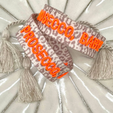 Close-up of adjustable woven friendship bracelet with Leo pattern and neon orange embroidered "PROSECCO, BABY!" lettering with sliding knot cords.