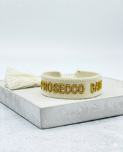 Close-up of adjustable ecru woven bracelet with gold embroidered “PROSECCO BABY” lettering and sliding knot tassel cords.