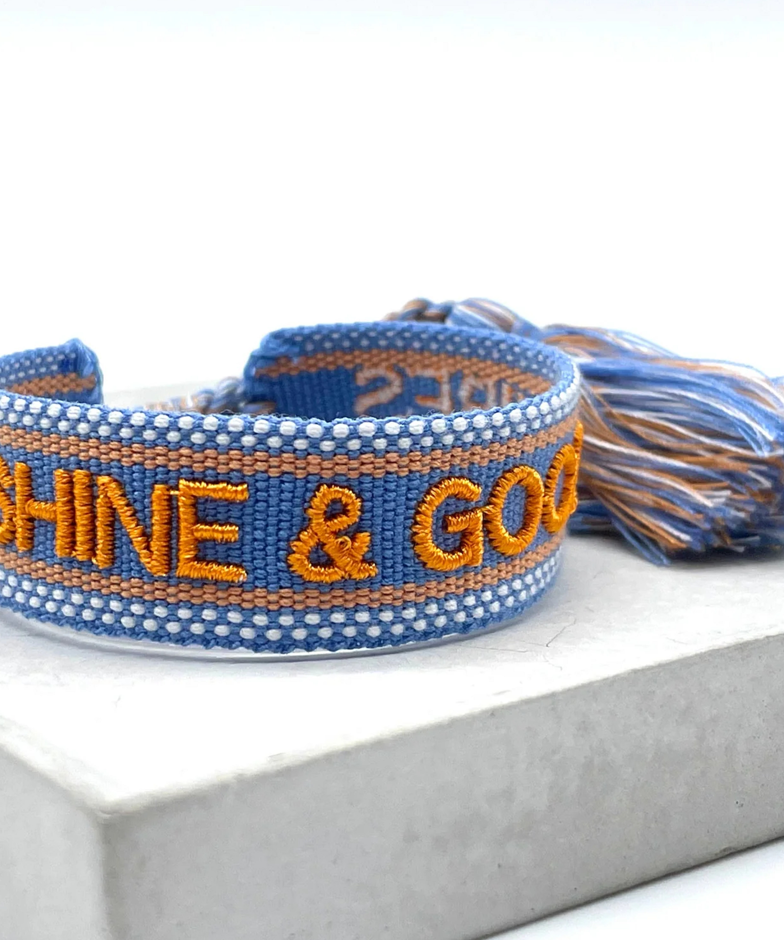 Close-up of adjustable blue and orange woven friendship bracelet featuring embroidered "SUNSHINE & GOOD VIBES" lettering and sliding knot tassel cords.