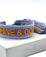 Close-up of adjustable blue and orange woven friendship bracelet featuring embroidered "SUNSHINE & GOOD VIBES" lettering and sliding knot tassel cords.