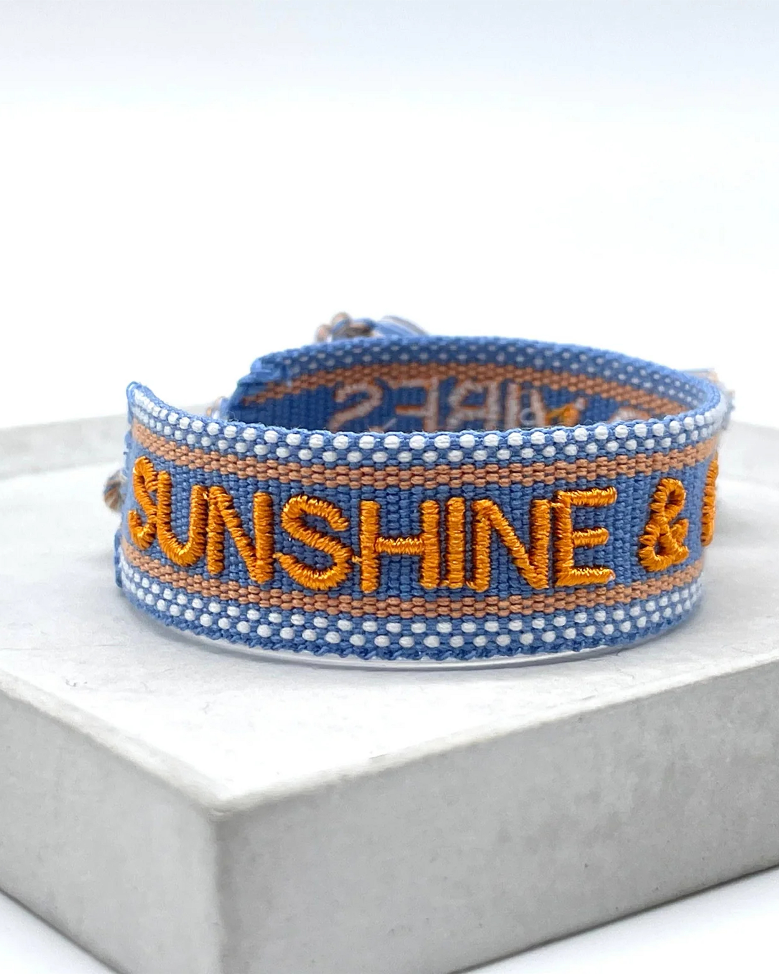 Close-up of adjustable blue and orange woven friendship bracelet featuring embroidered "SUNSHINE & GOOD VIBES" lettering and sliding knot tassel cords.