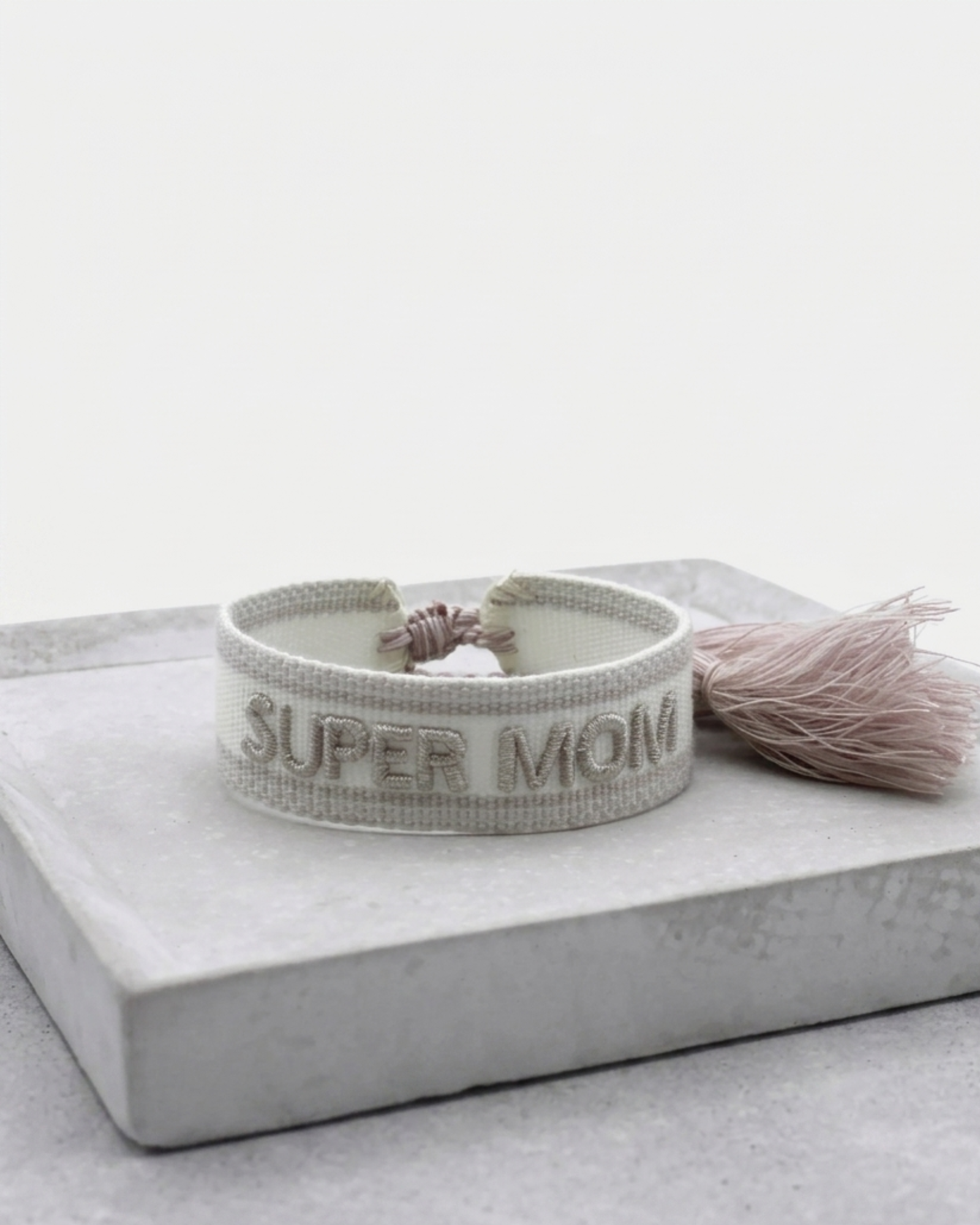 Close-up of adjustable white woven friendship bracelet with silver embroidered “SUPER MOM” lettering and sliding knot tassel cords.