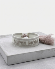 Close-up of adjustable white woven friendship bracelet with silver embroidered “SUPER MOM” lettering and sliding knot tassel cords.