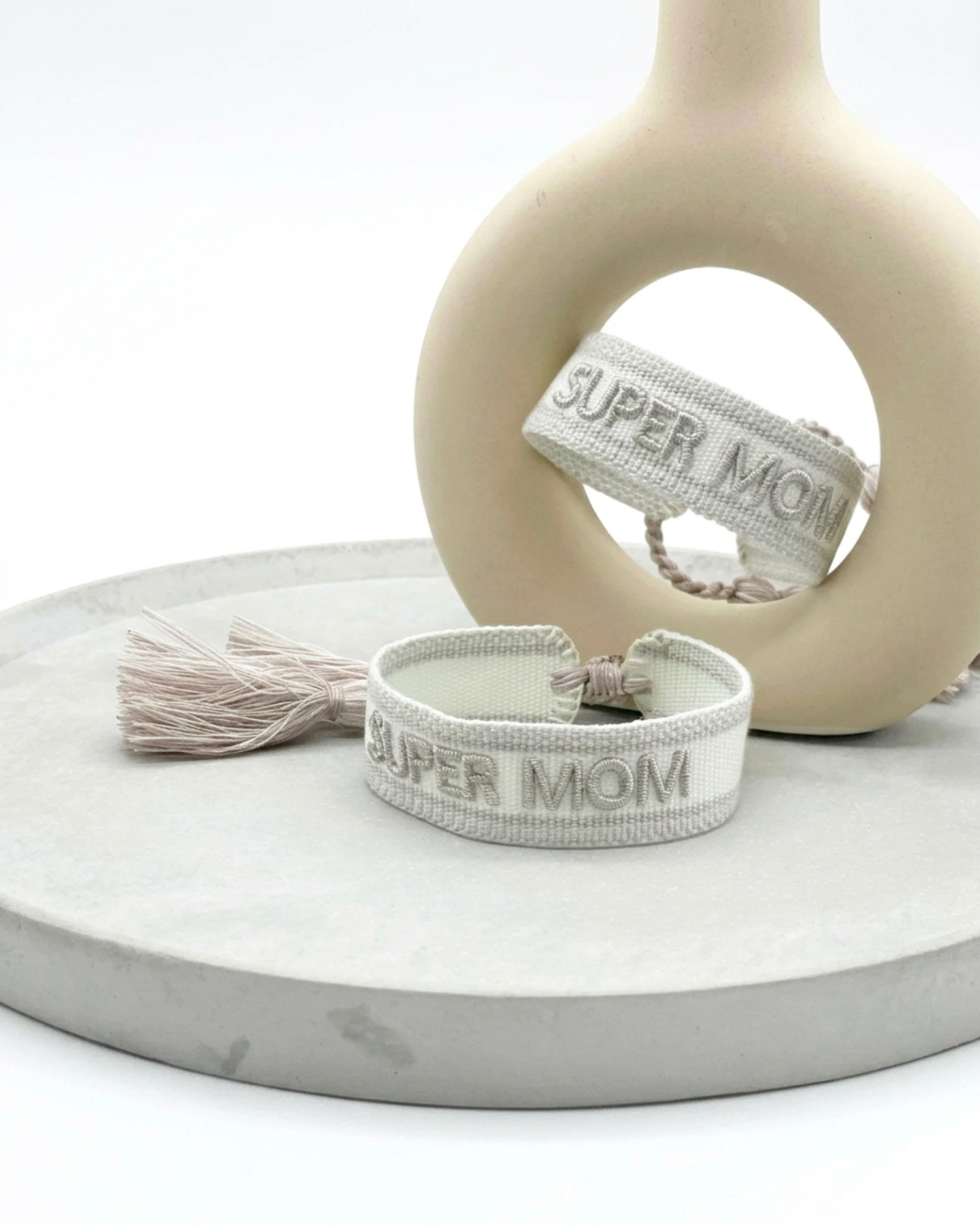 Close-up of adjustable white woven friendship bracelet with silver embroidered “SUPER MOM” lettering and sliding knot tassel cords.