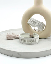 Close-up of adjustable white woven friendship bracelet with silver embroidered “SUPER MOM” lettering and sliding knot tassel cords.