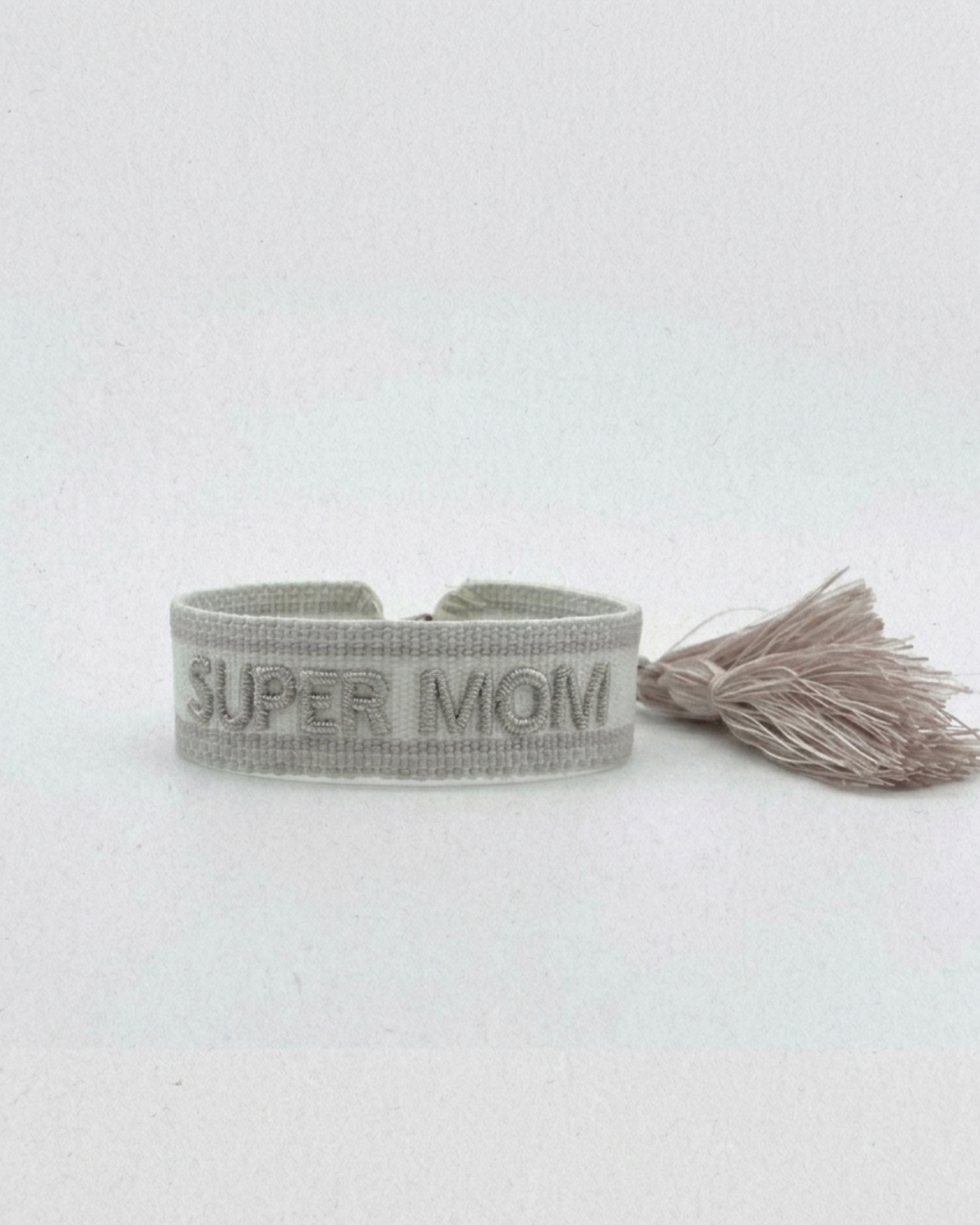 Close-up of adjustable white woven friendship bracelet with silver embroidered “SUPER MOM” lettering and sliding knot tassel cords.