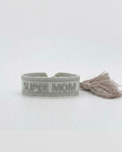 Close-up of adjustable white woven friendship bracelet with silver embroidered “SUPER MOM” lettering and sliding knot tassel cords.