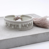 Close-up of adjustable white woven friendship bracelet with silver embroidered "SUPER MOM" lettering and sliding knot tassel cords.
