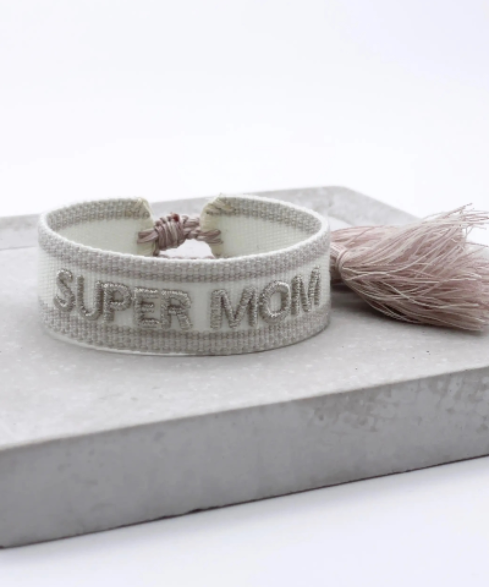 Close-up of adjustable white woven friendship bracelet with silver embroidered "SUPER MOM" lettering and sliding knot tassel cords.