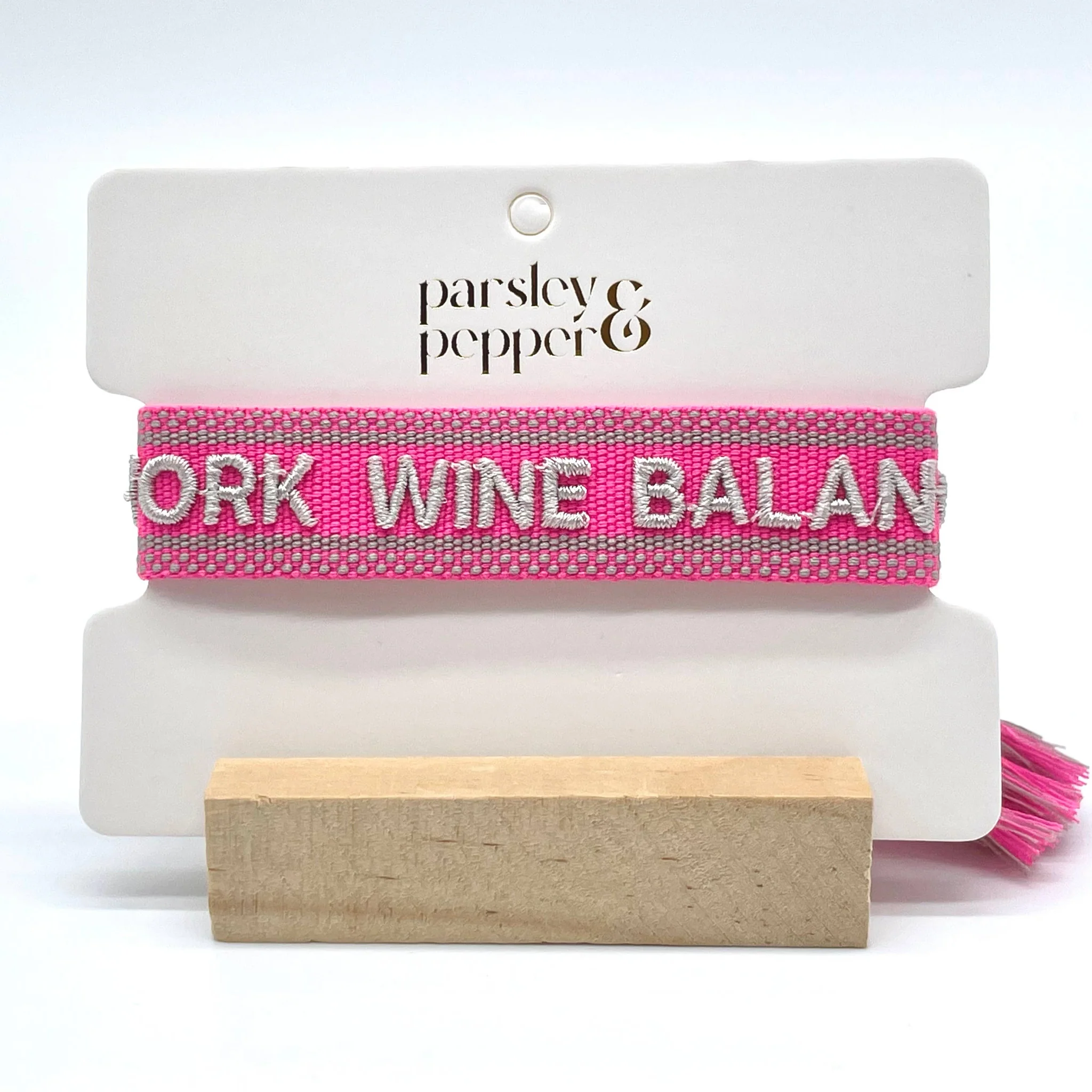 Close-up of adjustable pink woven bracelet with silver gray embroidered “WORK WINE BALANCE” lettering, matching border, and sliding knot tassel cords.
