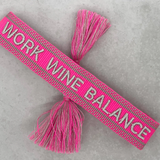 Close-up of adjustable pink woven bracelet with silver gray embroidered “WORK WINE BALANCE” lettering, matching border, and sliding knot tassel cords.