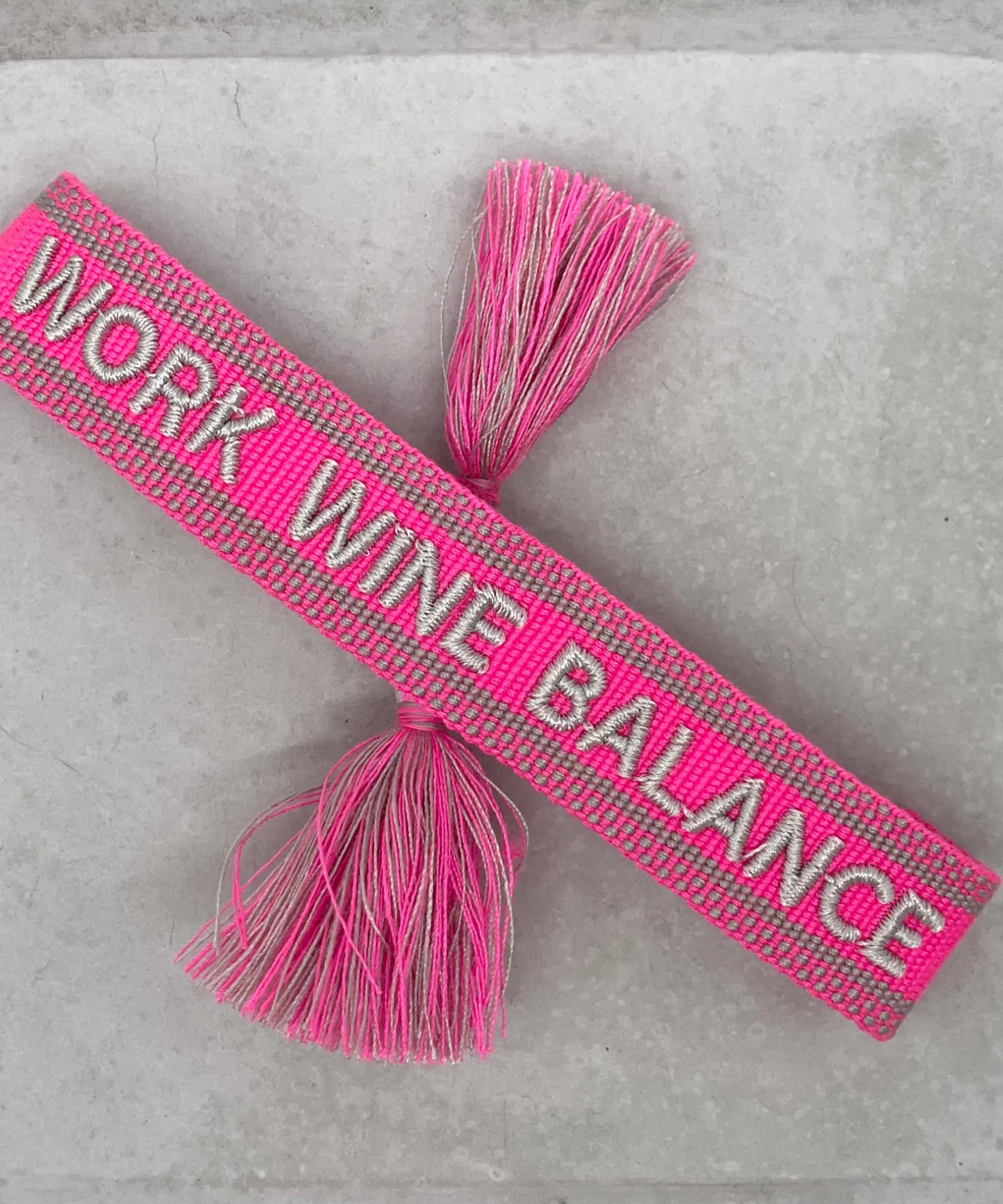 Close-up of adjustable pink woven bracelet with silver gray embroidered “WORK WINE BALANCE” lettering, matching border, and sliding knot tassel cords.