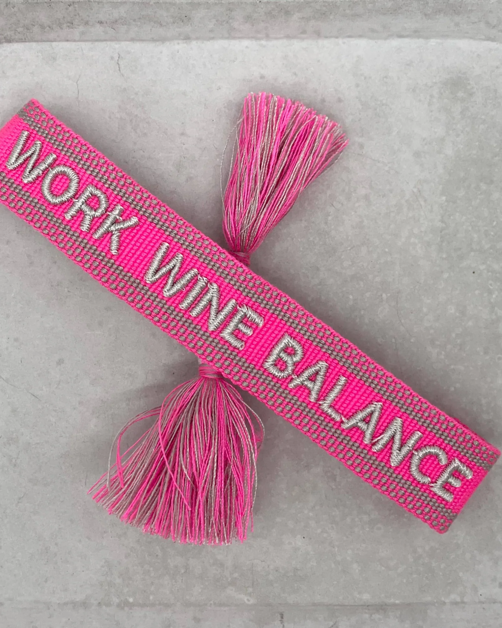 Close-up of adjustable pink woven bracelet with silver gray embroidered “WORK WINE BALANCE” lettering, matching border, and sliding knot tassel cords.