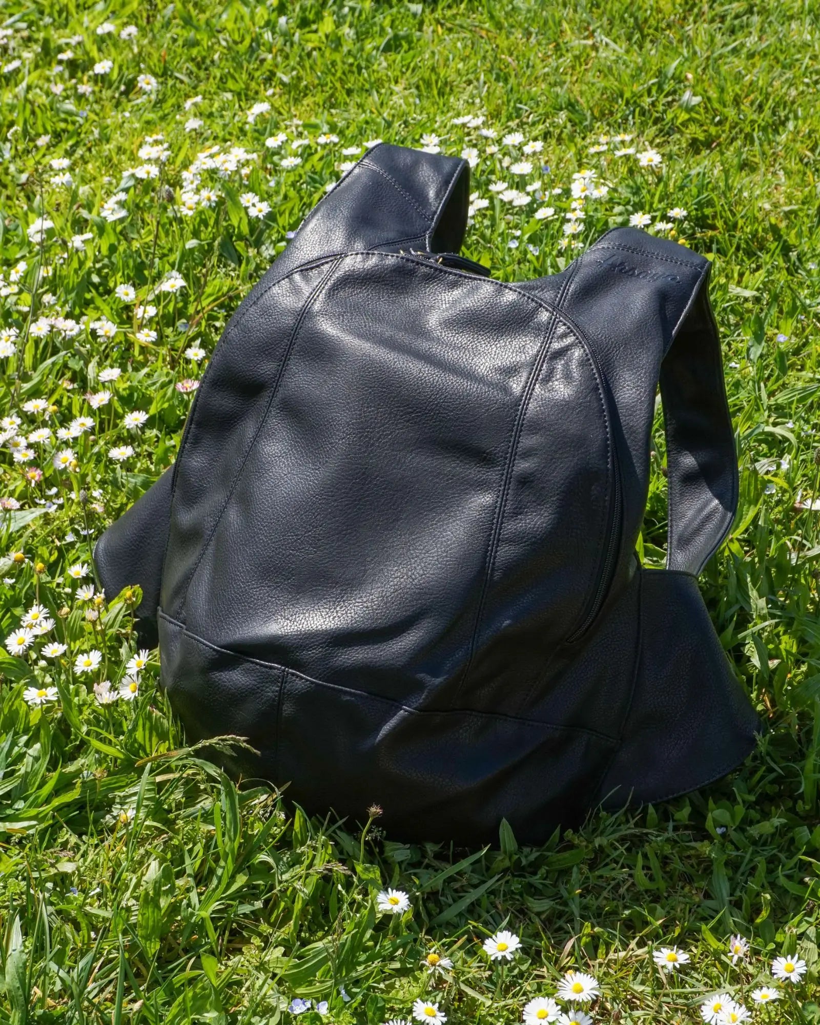 An Arsayo black vegan leather backpack sitting in a field of green grass and white wildflowers.