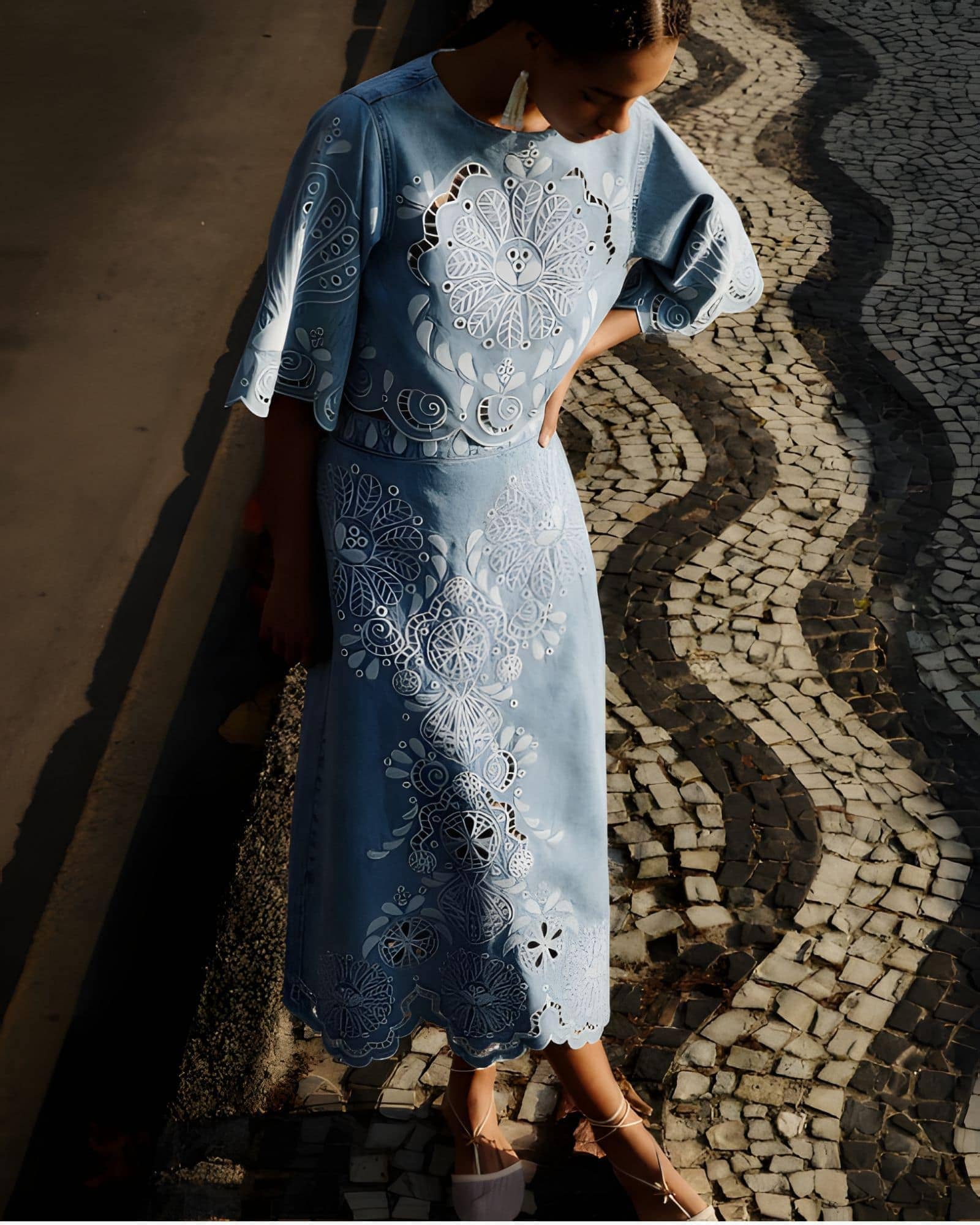 Full-length outdoor shot of a model wearing the Farm Rio denim Richelieu embroidered midi skirt on a patterned stone walkway.