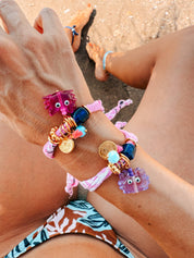 Colourful GOUPI bandana bracelet with a lilac glass crab charm on a person's wrist on summer vacations on a beach. 