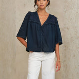 Model wearing Mabe Apparel navy embroidered cotton blouse with white trousers and jewelry.