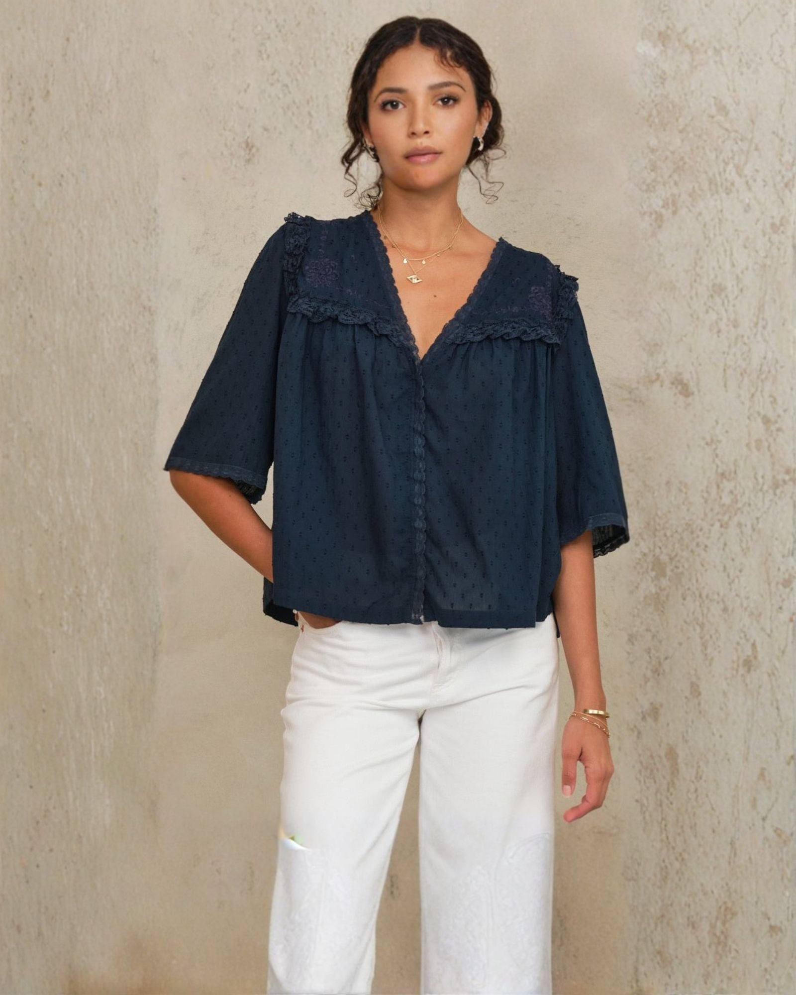 Model wearing Mabe Apparel navy embroidered cotton blouse with white trousers and jewelry.