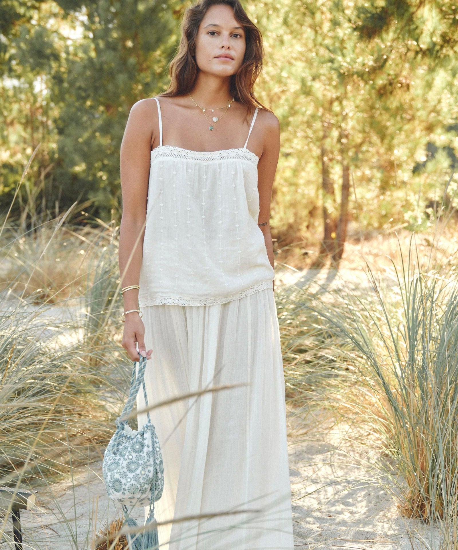 Woman in a Mabe Apparel cream cotton maxi skirt and matching camisole holding a woven bag outdoors.