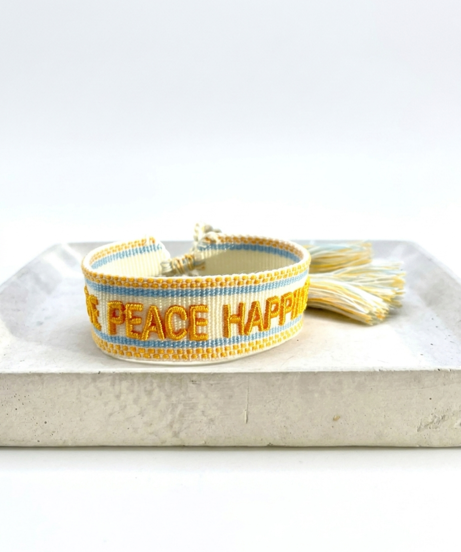 Cream woven Parsley & Pepper bracelet with mustard-yellow “LOVE PEACE HAPPINESS” embroidery, yellow and light blue borders and tasselled sliding knot.