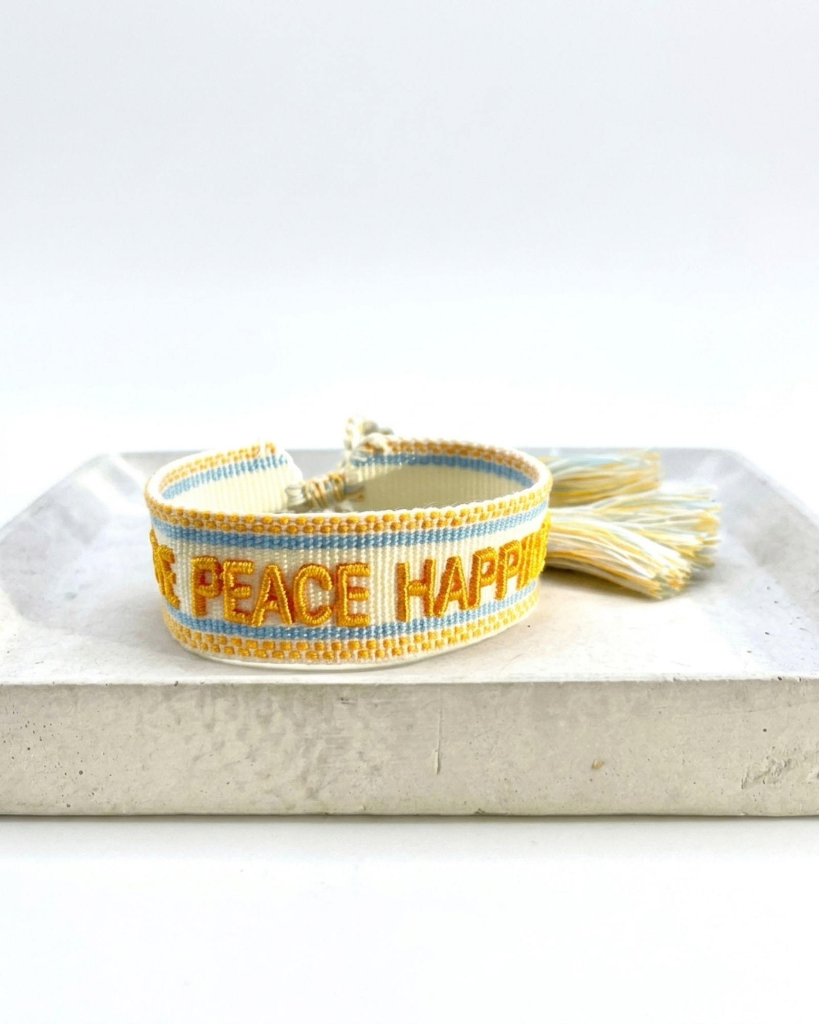 Cream woven Parsley & Pepper bracelet with mustard-yellow “LOVE PEACE HAPPINESS” embroidery, yellow and light blue borders and tasselled sliding knot.