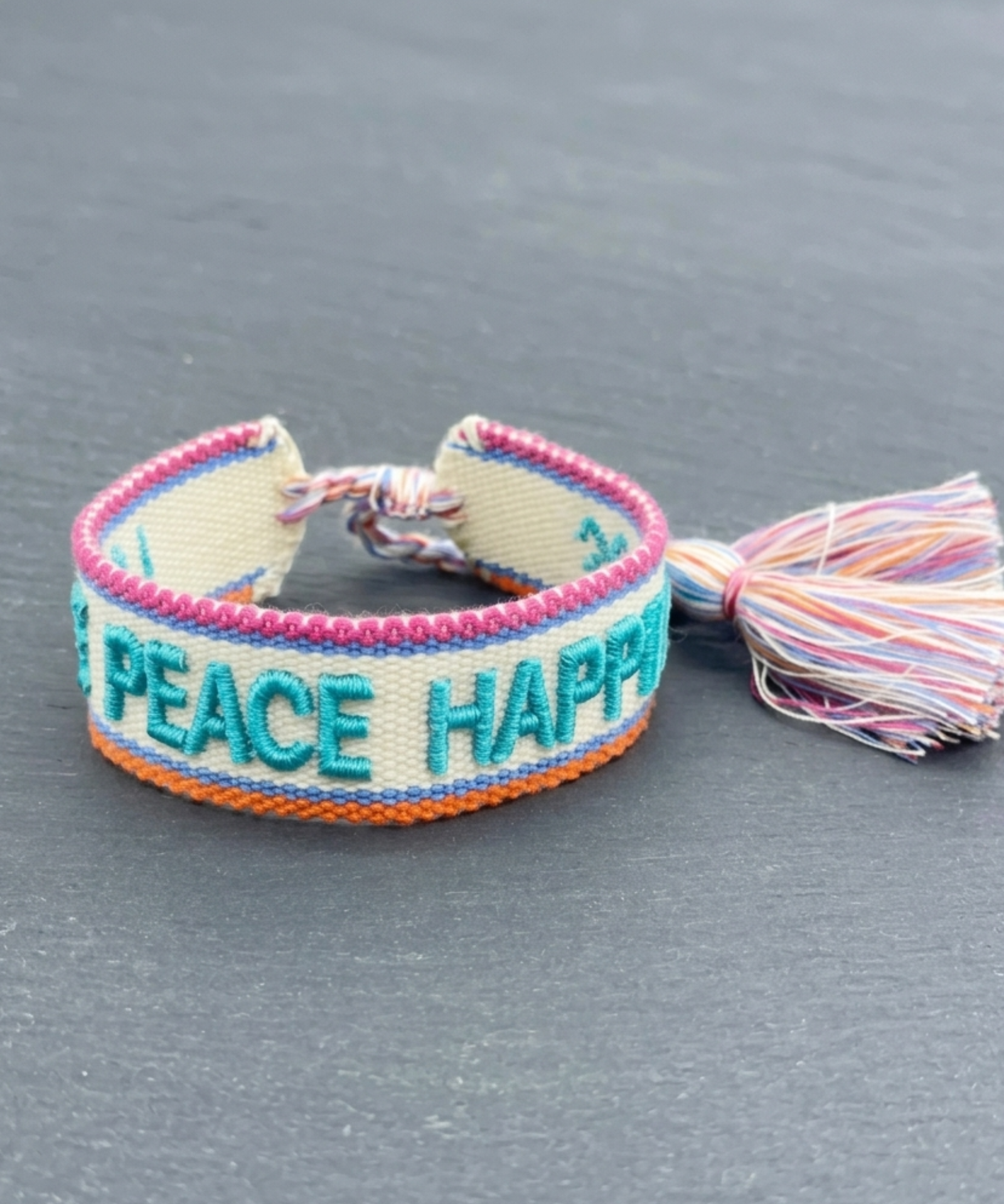Close-up of adjustable woven turquoise friendship bracelet with embroidered “LOVE PEACE HAPPINESS” lettering and sliding knot tassel cords.