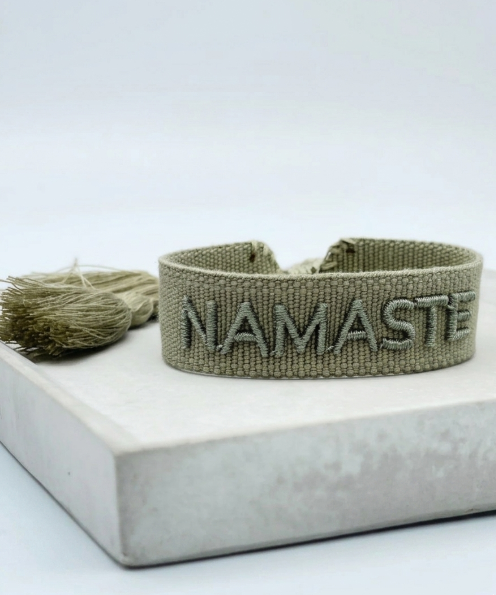 Close-up of adjustable sage eucalyptus woven bracelet with tone-on-tone embroidered “NAMASTE” lettering and sliding knot tassel cords.