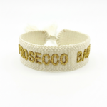 Close-up of adjustable ecru woven bracelet with gold embroidered “PROSECCO BABY” lettering and sliding knot tassel cords.