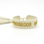 Close-up of adjustable ecru woven bracelet with gold embroidered “PROSECCO BABY” lettering and sliding knot tassel cords.