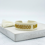 Close-up of adjustable ecru woven bracelet with gold embroidered “PROSECCO BABY” lettering and sliding knot tassel cords.